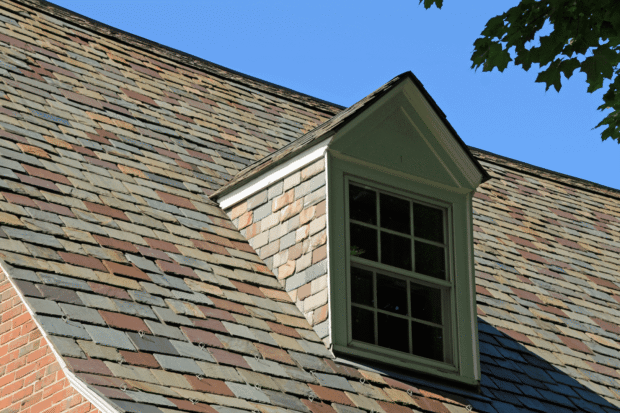 Slate roof with various colors, featuring a dormer window, against a clear blue sky. Green window trim contrasts with the shingles. Tree leaves partially visible in the top right corner.